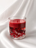 A clear glass candle vessel filled with deep red wax and red botanical elements, set against a draped white fabric background.