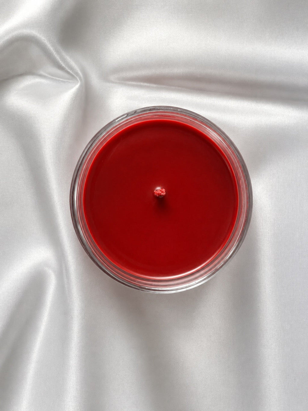 A top-down view of a round red candle in a clear glass container, set against a draped white satin fabric background.