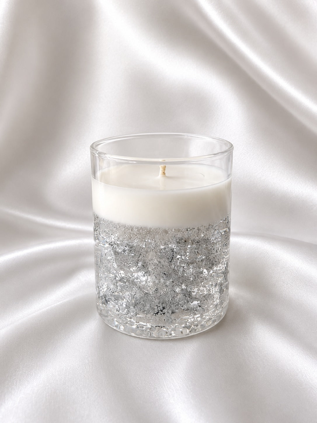 A glass jar candle with a white wax top and a bottom half filled with silver glitter, set against a draped white satin fabric background.