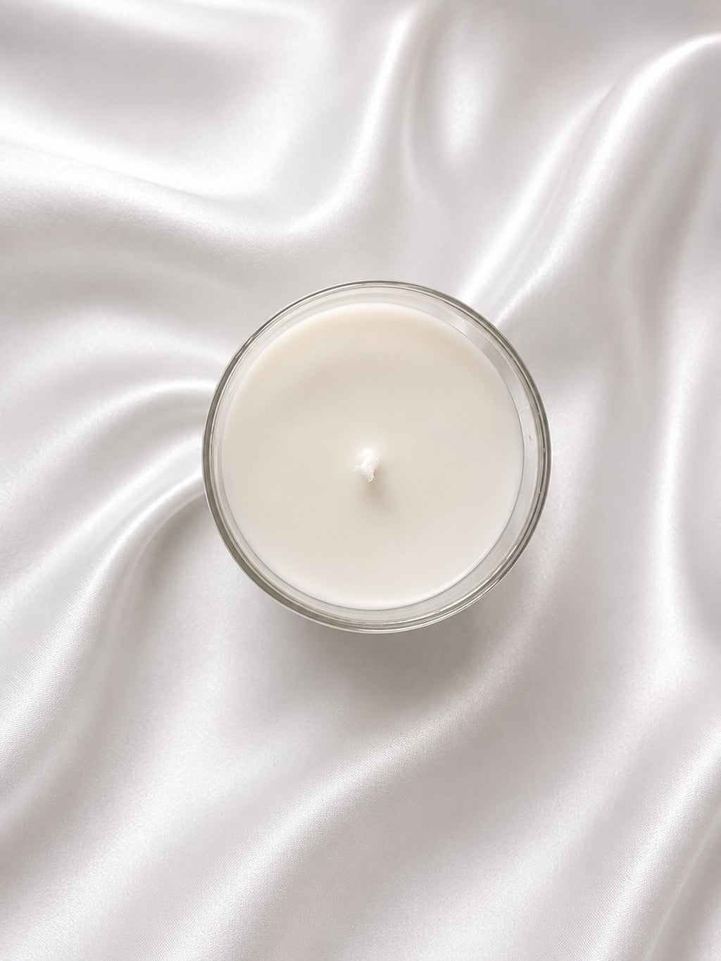 A single-wick candle in a clear glass jar with creamy off-white wax, centered on draped white satin fabric.