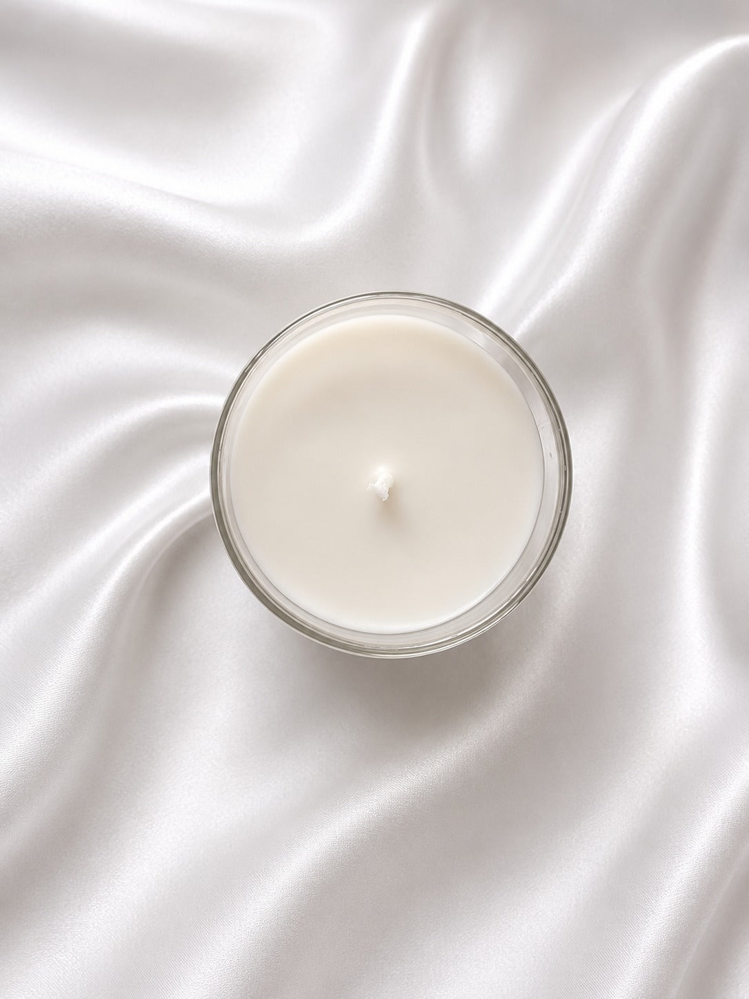 A single-wick candle in a clear glass jar with creamy off-white wax, centered on draped white satin fabric.