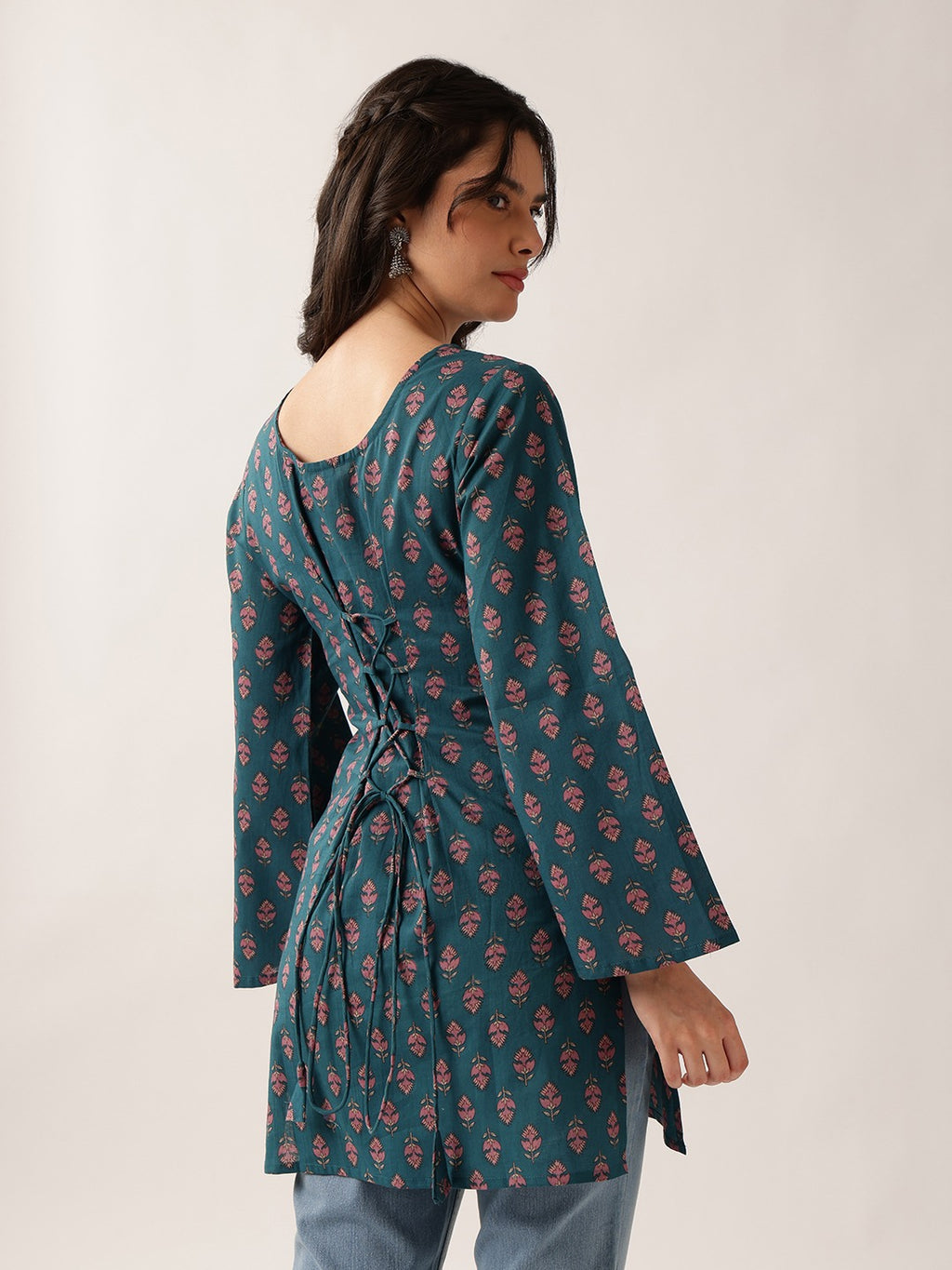 A woman wearing a teal green long-sleeve tunic with a pink and red floral block print, shown from a three-quarter back view against a plain light beige studio background.