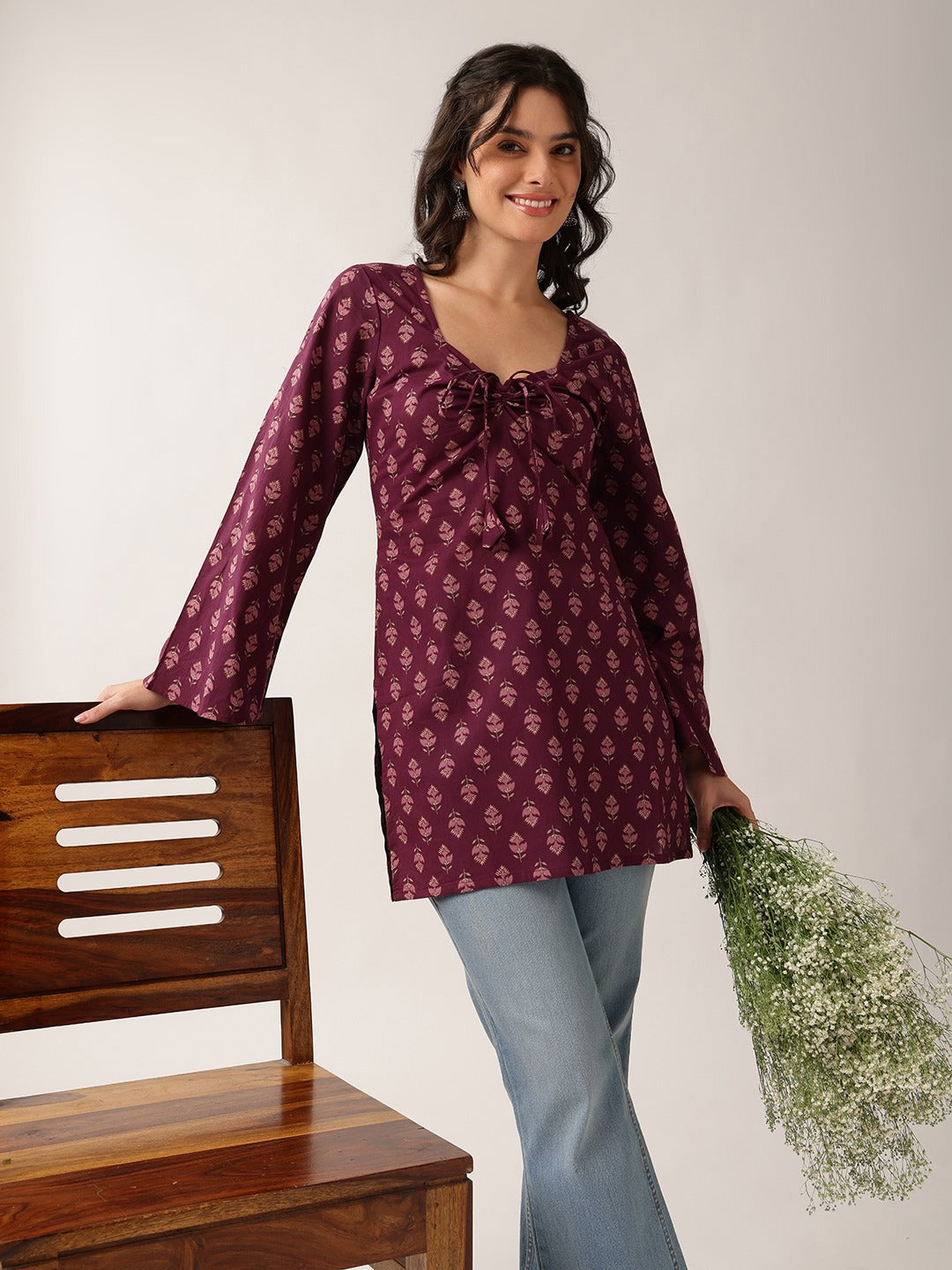 A woman wearing a long-sleeve maroon tunic top with a small white floral print and a drawstring neckline, paired with light blue jeans, standing next to a wooden chair and holding a bunch of green baby's breath.