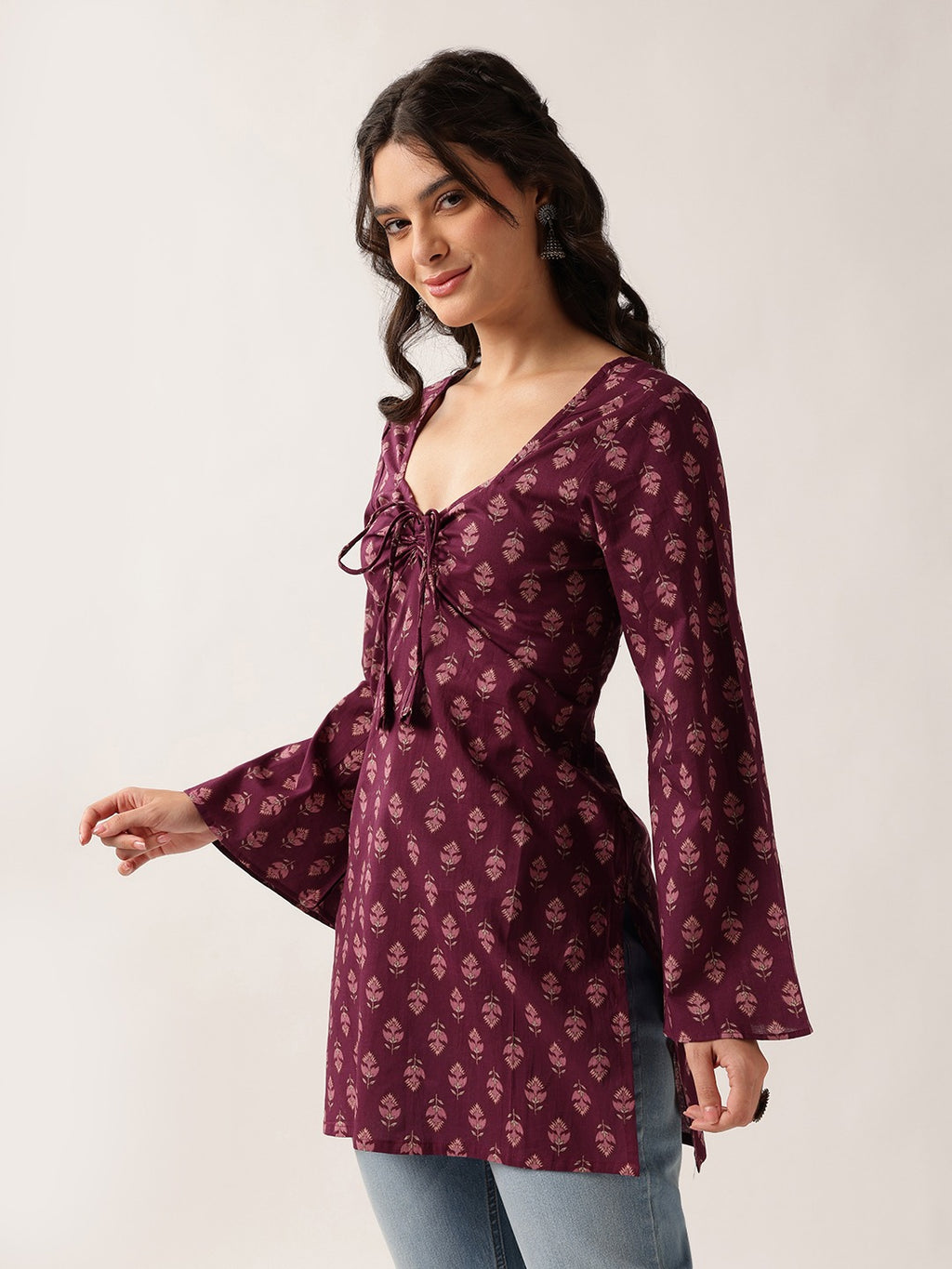 A woman modeling a long-sleeve maroon tunic top with a small floral block print and a front tie detail, paired with light blue jeans against a plain light gray background.