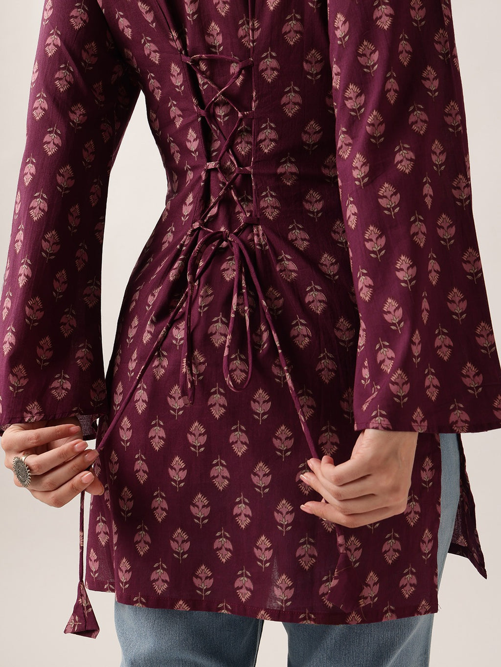 A close-up of a person wearing a long-sleeve maroon tunic with a beige floral block print and a lace-up back detail, paired with blue jeans.