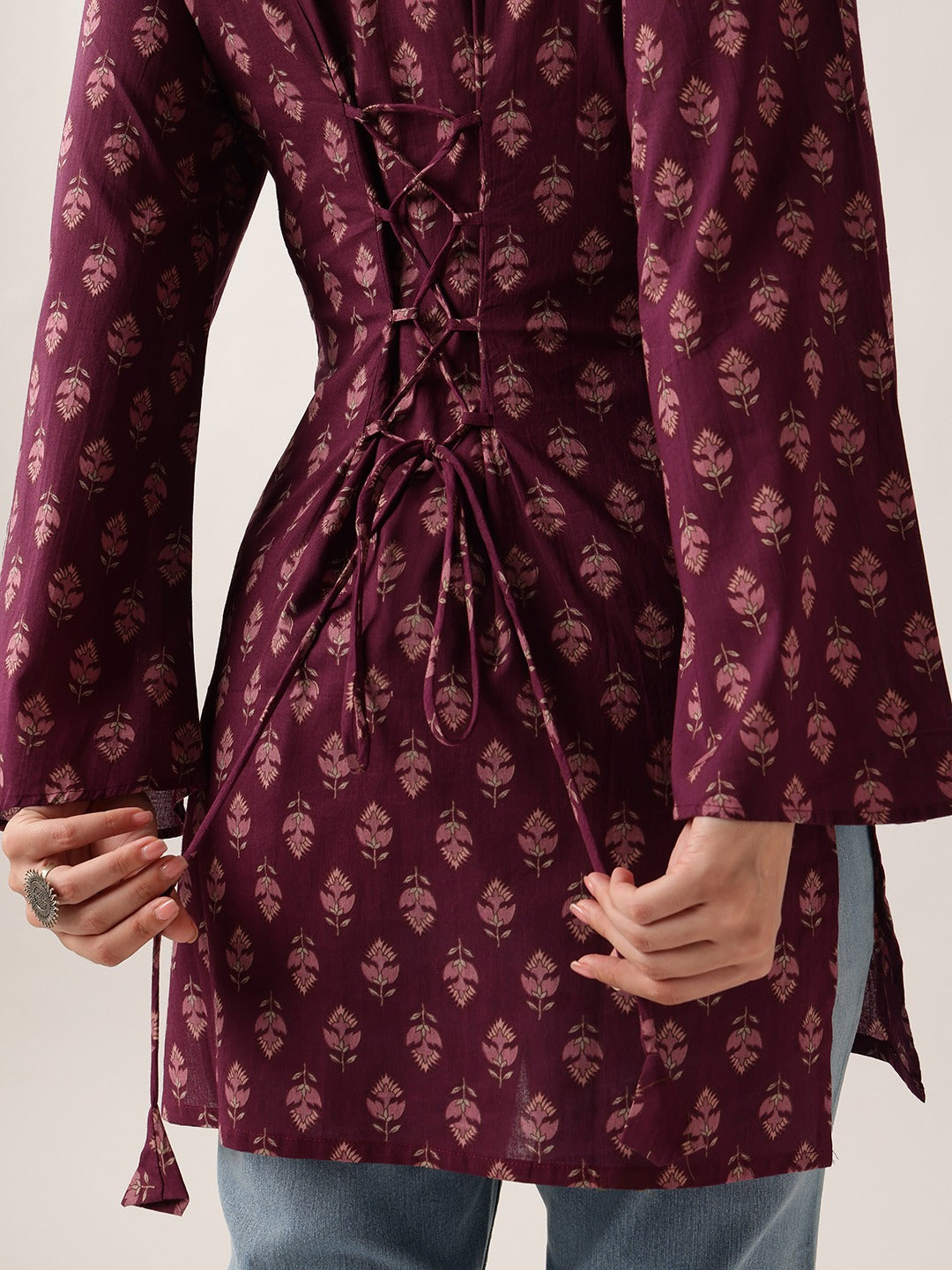 A close-up of a person wearing a long-sleeve maroon tunic with a beige floral block print and a lace-up back detail, paired with blue jeans.