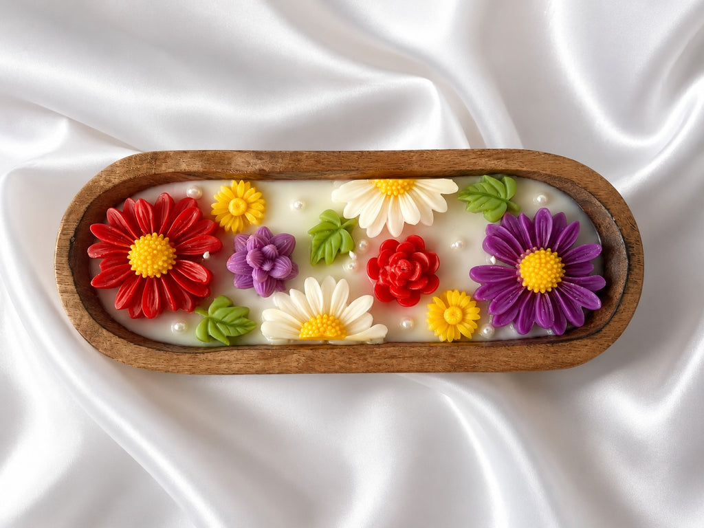 An oval wooden tray containing a white wax melt or soap base decorated with colorful sculpted flowers in red, purple, yellow, and white, with green leaves, set against a draped white satin background.