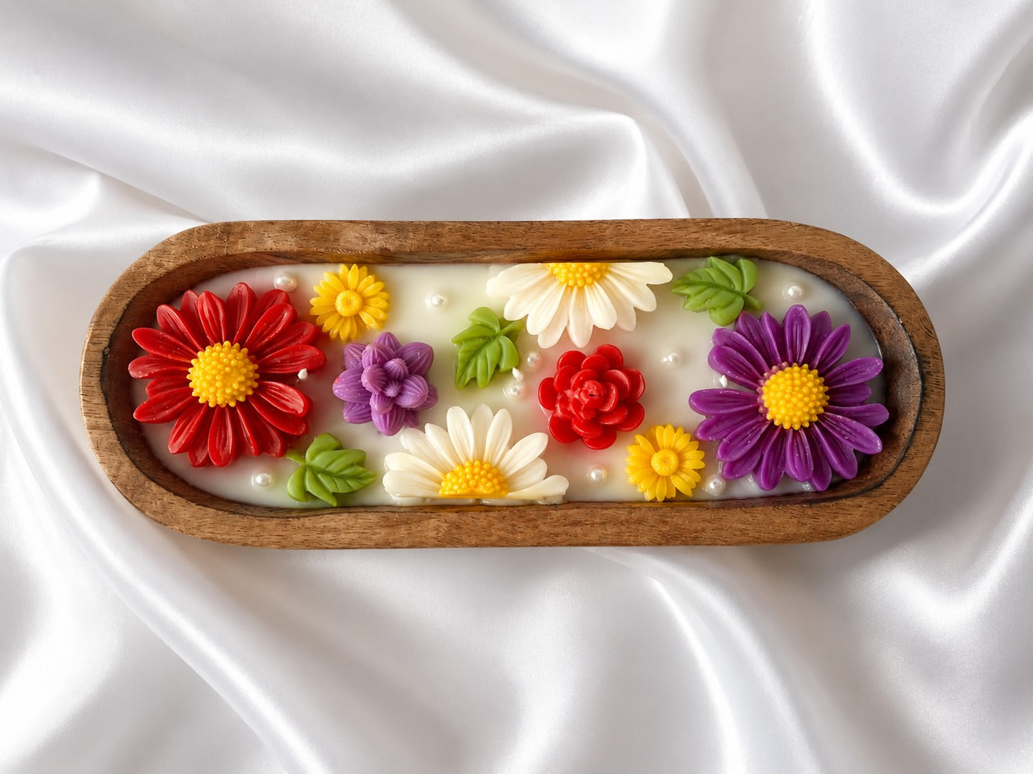 An oval wooden tray containing a white wax melt or soap base decorated with colorful sculpted flowers in red, purple, yellow, and white, with green leaves, set against a draped white satin background.