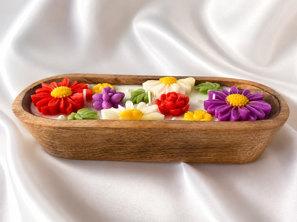 A decorative oval wooden tray filled with colorful flower-shaped candles or wax ornaments in red, purple, white, and yellow, set against a draped white satin background.