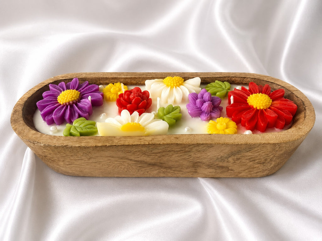 A wooden oval tray filled with white wax melts or soap flakes, decorated with colorful sculpted flowers in purple, red, white, yellow, and green, set against a draped white satin background.