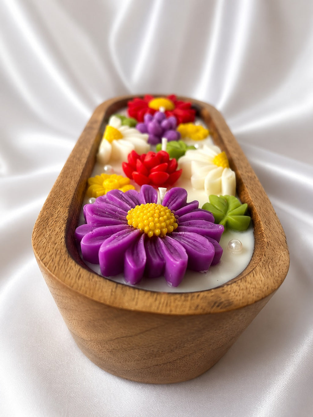 An oval wooden candle holder filled with white wax and decorated with colorful sculpted flowers in purple, red, yellow, and green, set against a draped white satin background.