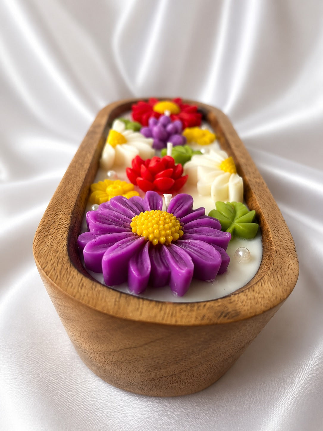 An oval wooden candle holder filled with white wax and decorated with colorful sculpted flowers in purple, red, yellow, and green, set against a draped white satin background.